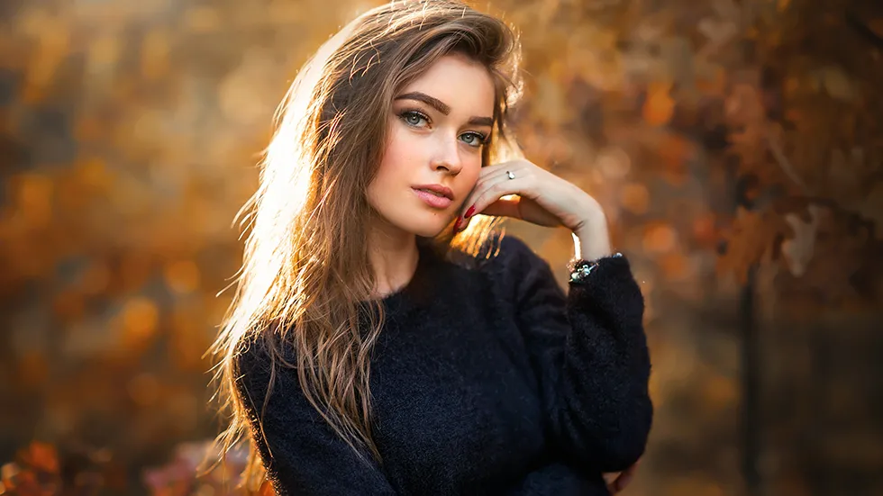 A young woman with long brown hair looks at the camera while standing in front of a blurred background of fall leaves.